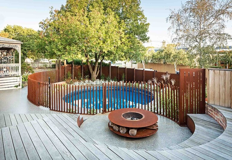 Circular blue pool surrounded by curved corten steel slat fencing, timber decking and central fire pit in Hawthorn backyard