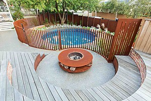 Curved timber decking surrounds corten steel fire pit with circular pool and vertical screening in Hawthorn garden