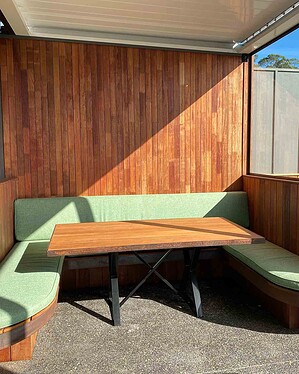 Built-in corner banquette with green upholstery and timber table under covered outdoor area in Gisborne garden