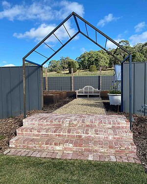 Steel archway frame over brick steps leading to enclosed garden courtyard with bench and plantings in Gisborne