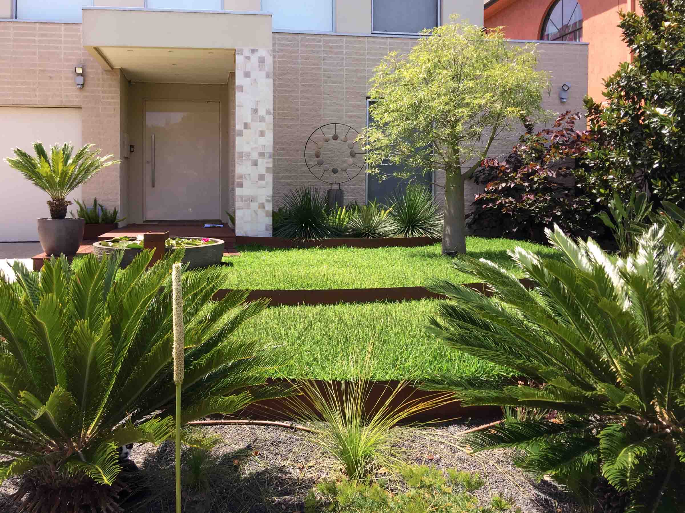 Modern front garden with stepped lawn levels, native plants, and contemporary house entrance with beige stone cladding